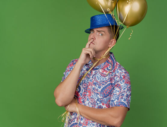 抱着一个体贴的年轻人 戴着蓝色帽子 脖子上绑着气球 手指放在脸上 脸上贴着绿色帽子脸颊领带