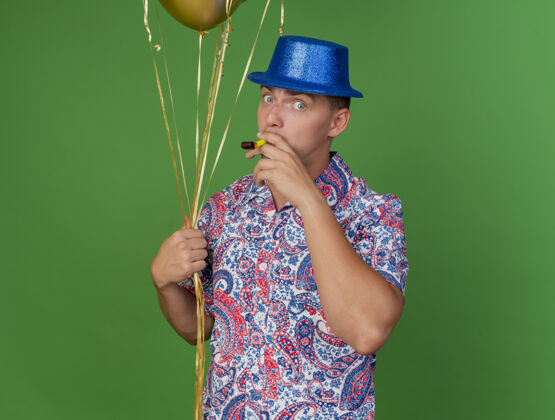 年轻人高兴的年轻人戴着蓝帽子 手持气球 吹着绿色的派对风机蓝色小伙子绿色
