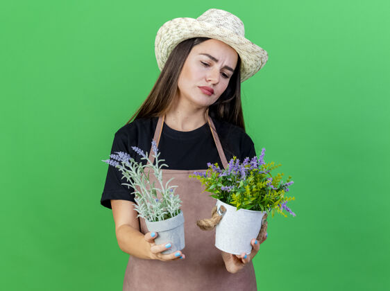 园艺体贴美丽的园丁女孩穿着制服戴着园艺帽捧着花盆望着隔离在绿色上的花朵穿帽子制服
