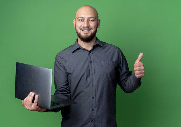 脸微笑的年轻秃头呼叫中心男子手持笔记本电脑 并显示拇指向上孤立的绿色墙上公民人姿势