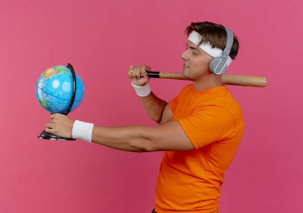 个人资料高兴的年轻英俊的运动型男子戴着头带和腕带 戴着耳机站在侧视图肩上拿着棒球棒和地球仪 看着隔离在粉红色墙上的地球仪站着运动帅气