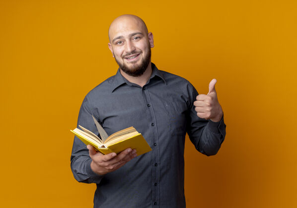 人微笑着的年轻秃头呼叫中心的男人拿着一本书 在橙色的墙上孤立地竖起大拇指人脸人