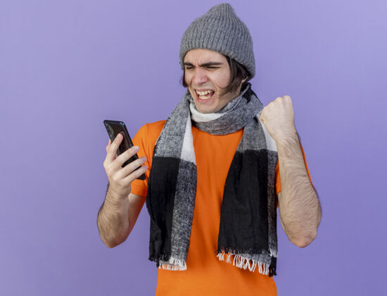 紫色快乐的年轻病夫戴着冬天的帽子 戴着围巾 看着手机 在紫色上显示“是”的手势年轻手势拿着