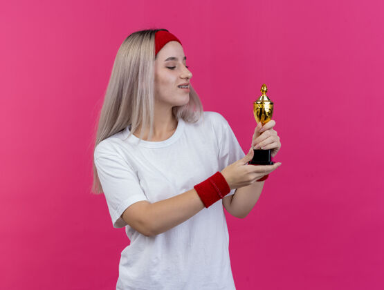 腕带戴着头带和腕带的戴着牙套的快乐的年轻的运动女性拿着并看着隔离在粉红色墙上的冠军杯女孩运动型头带
