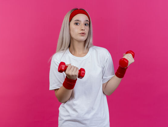 女人令人印象深刻的年轻运动女性戴着背带 戴着头带和腕带 举着孤立在粉红色墙上的哑铃人女孩印象