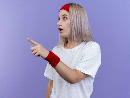 壁板惊讶的年轻运动女性戴着背带 戴着头带和腕带 看着紫色墙上孤立的一面点人女人
