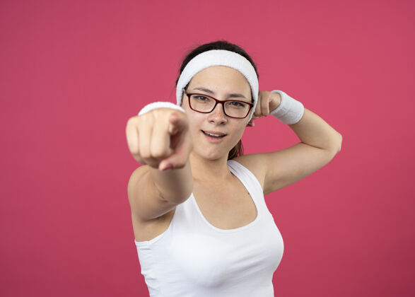 紧张自信的年轻运动女性戴着眼镜戴着头带和腕带紧张的二头肌和点在前面隔离在粉红色的墙上头带光学信心