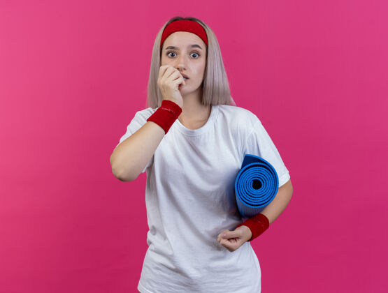 头带焦虑的年轻运动女性戴着背带 戴着头带和腕带 咬着指甲 拿着运动垫子孤立地站在粉红色的墙上女人人背带