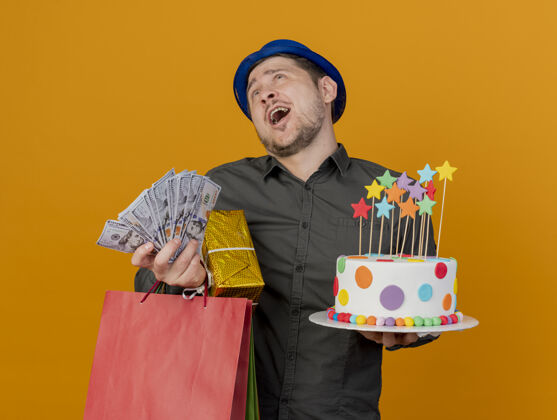 举行快乐的年轻人戴着蓝色的帽子拿着蛋糕和礼物和钱孤立在橙色钱小伙子穿