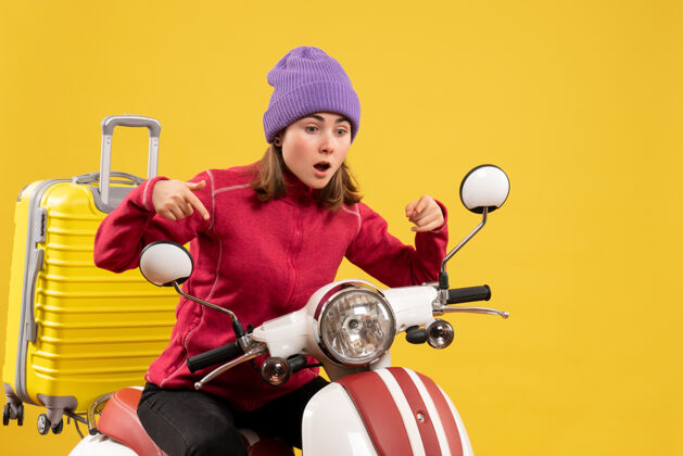 前面前视图困惑的坐在轻便摩托车上的年轻女子指着什么东西指向快乐帽子