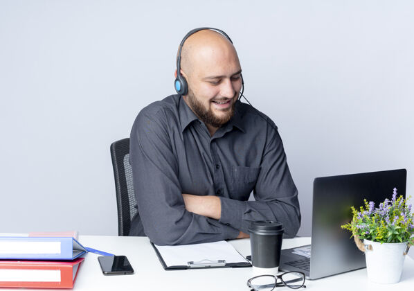 电话微笑着的年轻秃头呼叫中心男子戴着耳机坐在办公桌旁 紧闭的姿势拿着工作工具 看着隔离在白墙上的笔记本电脑微笑姿势人
