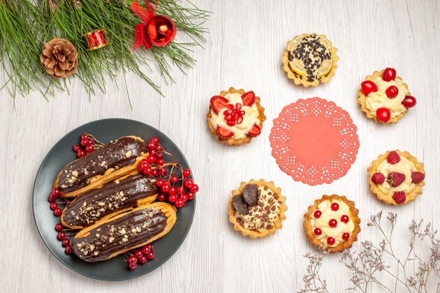 晚餐上图：灰色盘子上的巧克力蛋糕和醋栗红色椭圆形蕾丝小饰物 圆形的馅饼和松树叶子 白色木质地面上有圣诞玩具花边木头椭圆形