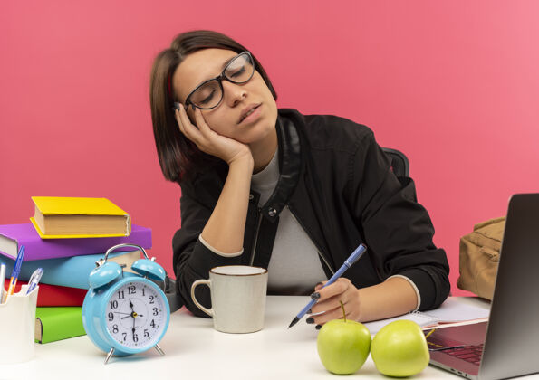 人昏昏欲睡的年轻女学生戴着眼镜坐在书桌旁 拿着钢笔 手放在脸上 闭着眼睛做作业 隔离在粉红色的墙上姿势脸眼睛