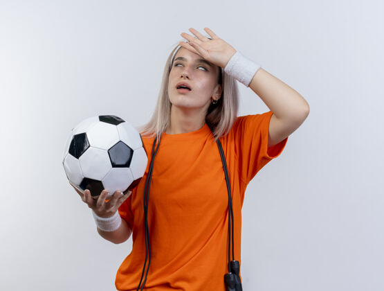 人戴着牙套 脖子上系着跳绳 戴着头带和腕带 拿着球 把手放在隔离在白墙上的额头上的年轻运动型女人球背带脸