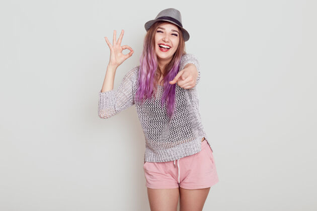 享受紫色头发的兴奋快乐的女人用手指着相机 表示同意的手势 表达积极的情绪 隔着白色的墙好指指点点选择