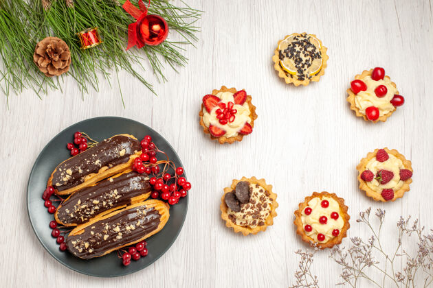 酸顶视图巧克力eclairs和醋栗在灰色板馅饼和松树叶子与圣诞玩具在白色的木制地面玩具晚餐食物
