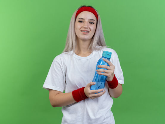 微笑微笑的年轻运动女性戴着背带 戴着头带和腕带 把水瓶隔离在绿色的墙上持有穿人