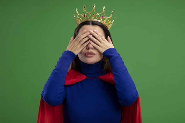 穿高兴的年轻超级英雄女孩戴着皇冠戴着眼睛 双手孤立在绿色封面超级英雄王冠