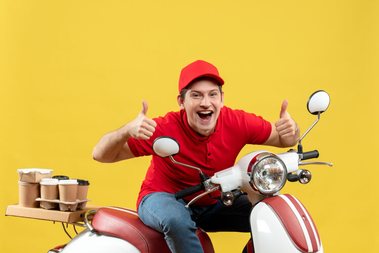 年轻人俯视图：微笑的年轻人穿着红色上衣 戴着帽子 坐在踏板车上 在黄色的墙上做着“ok”的手势 传递着命令人运动滑板车
