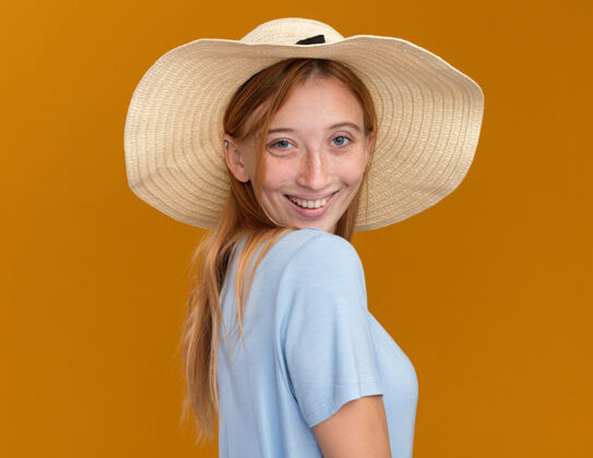 站着带着雀斑的年轻红发姜红色女孩 戴着沙滩帽 站在橙色的沙滩上 面带微笑红发帽子微笑