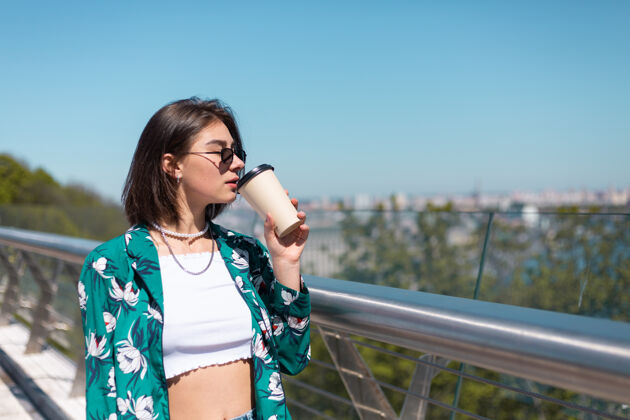 积极户外写真：穿着绿衬衫的女人 喝着咖啡 享受着阳光 站在桥上 清晨的城市景色令人惊叹城镇姿势早晨
