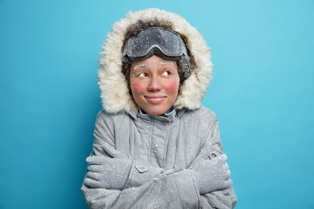 室内冻僵的女人拥抱着自己 浑身冻得发抖 满脸白霜 神情愉快地戴着滑雪板护目镜 冬衣和手套 愉快地微笑着可爱兜帽女性