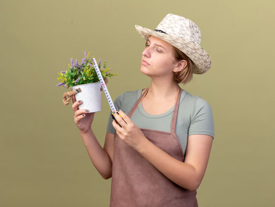 措施自信的年轻斯拉夫女园丁戴着园艺帽在橄榄绿上用卷尺量花盆斯拉夫胶带花园
