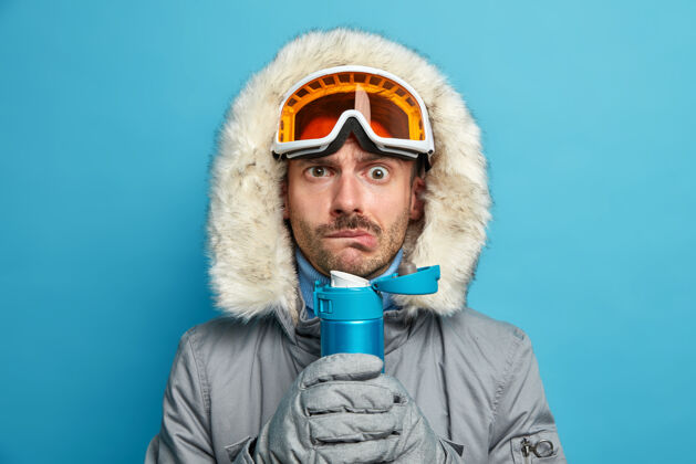 严肃严肃的男人在寒冷的冬天滑雪后会因寒冷而发抖 拿着装有热饮料的瓶子 戴着滑雪镜和暖和的夹克娱乐颤抖滑雪