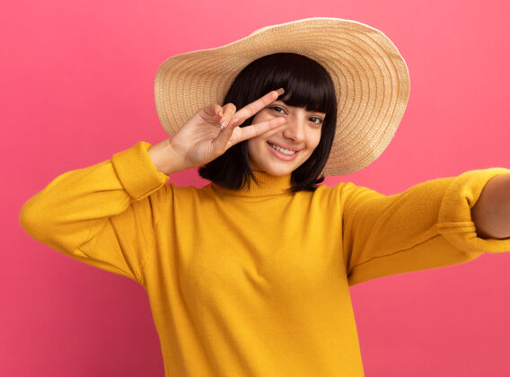 海滩微笑着的年轻黑发白人女孩戴着沙滩帽手势胜利标志在粉红色上自拍姿态黑发胜利