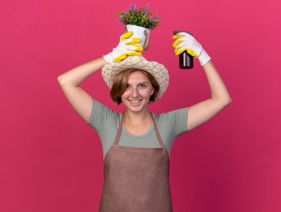 瓶微笑着的年轻斯拉夫女园丁戴着园艺帽和手套拿着喷雾瓶和花盆在粉红色的头上帽子女年轻