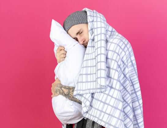 年轻人疼痛的年轻白种人 戴着冬天用格子布包裹的帽子 抱着他 把头放在枕头上 隔离在粉红色的墙上 留着复印空间拥抱头帽子