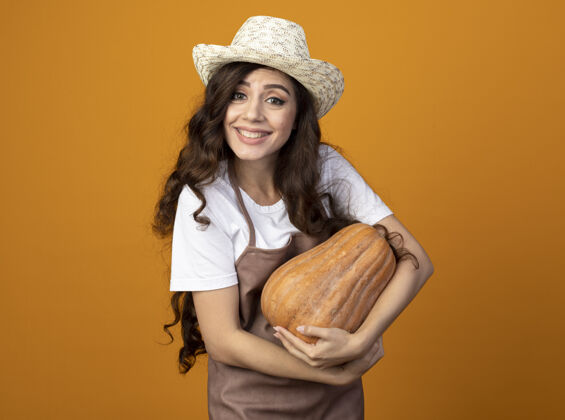 年轻微笑的年轻女园丁穿着制服 戴着园艺帽 手里拿着南瓜 隔离在橙色的墙上 留着复制空间穿帽子南瓜