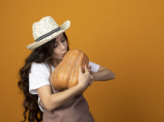 穿高兴的年轻女园丁穿着制服戴着园艺帽假装亲吻孤立在橙色墙上的南瓜复制空间园艺制服南瓜