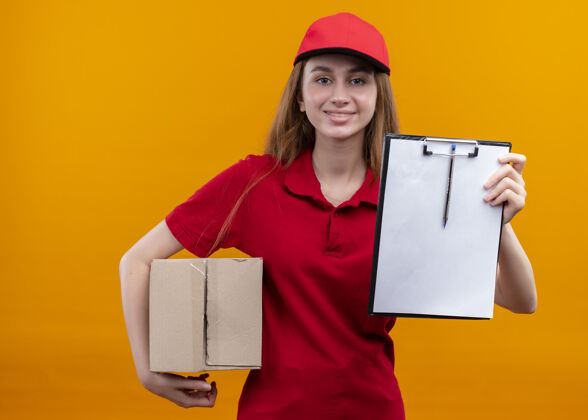 盒子微笑着的年轻送货女孩 穿着红色制服 拿着盒子 在孤立的橙色空间里展示着剪贴板女孩剪贴板交货