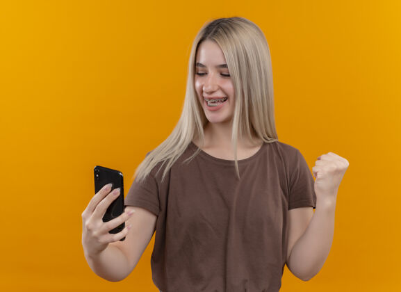 电话戴着牙套的快乐的金发女孩手持手机 在孤立的橙色空间里举起拳头看着它持有牙套金发