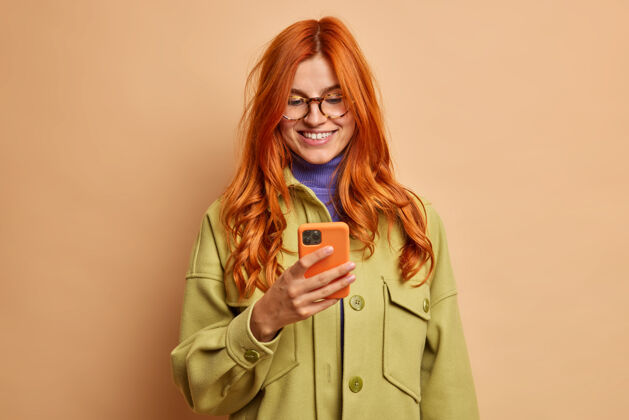 魅力红发美女笑容可掬地使用手机应用程序很高兴收到男友的短信穿着时髦的秋装在网上进行愉快的交谈欢呼单身红发