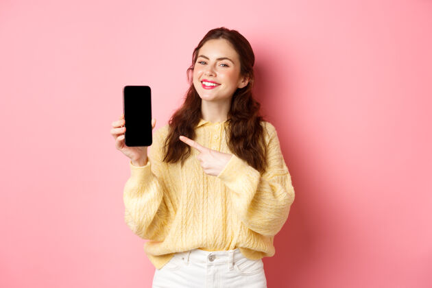 科技一位年轻漂亮的女士站在粉红色的墙上 用手指着智能手机 微笑着 在手机上演示宣传片积极情感在线