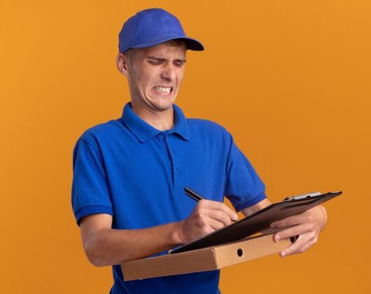 盒子恼怒的年轻金发送货员拿着笔写在剪贴板上的比萨饼盒上的橙色金发橘子钢笔