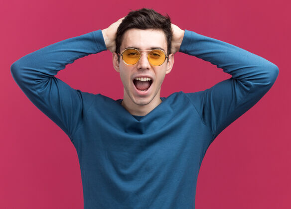 相机快乐的年轻黑发白人男孩戴着太阳眼镜双手放在头上看着粉红色的相机眼镜粉色快乐