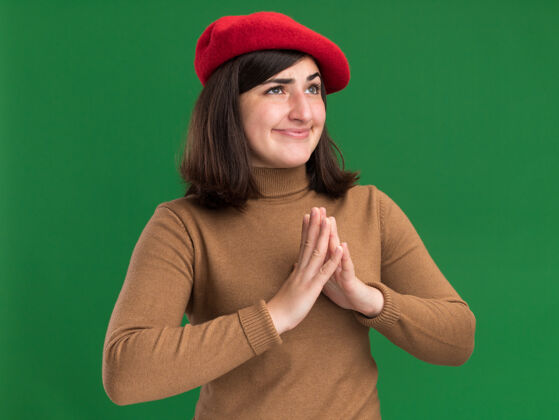 帽子邪恶快乐的年轻漂亮的高加索女孩戴着贝雷帽 双手合十 看着绿色的一面邪恶年轻壁板