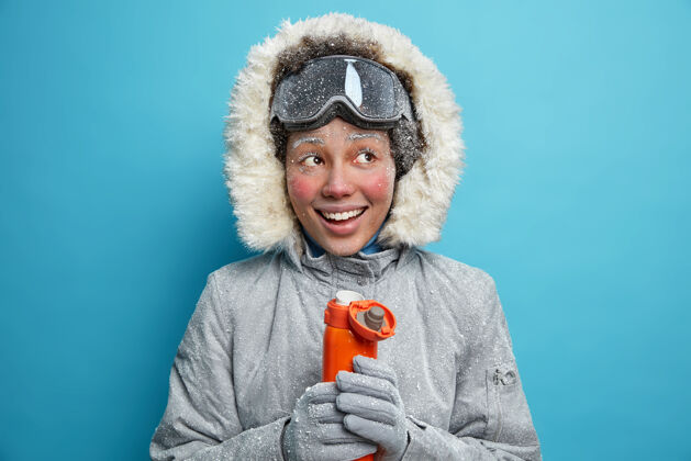 乐趣穿着冬装的快乐霜冻女人在寒冷的天气里用热水瓶里的热茶取暖微笑着宽大地戴着滑雪镜积极地休息勇敢积极的女远足者寒冷年轻滑雪