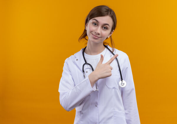 穿高兴的年轻女医生穿着医用长袍和听诊器指着右边的橙色空间和复印空间指向医生右
