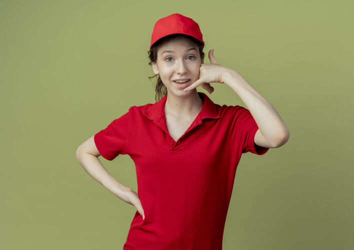 手势令人印象深刻的年轻漂亮的送货女孩在红色制服和帽子把手放在腰上 并在橄榄绿的背景与复制空间孤立做呼叫手势腰手背景