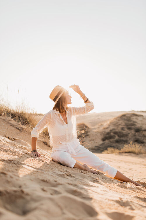 旅行穿着白色套装 戴着草帽的沙漠沙滩上的时髦美女体验热沙漠