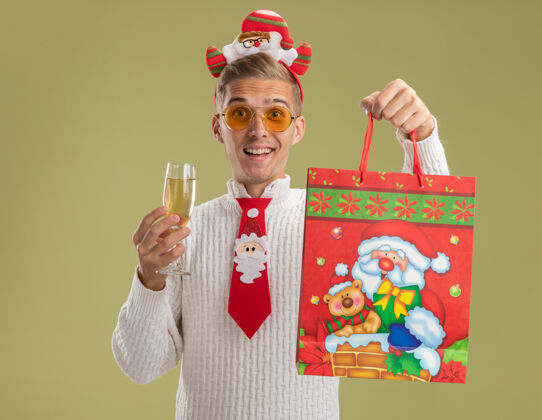 杯子印象深刻的年轻帅哥戴着圣诞老人的头带和领带看着相机拿着一杯香槟和圣诞礼包孤立在橄榄绿的背景圣诞老人绿色礼物