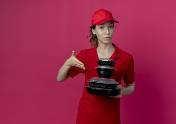 持有令人印象深刻的年轻漂亮的送货女孩穿着红色制服和帽子举行 并用手指着食品容器隔离在深红色的背景与复制空间制服印象手