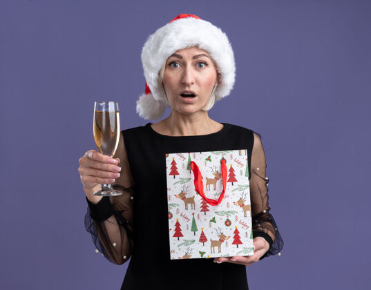 帽子印象深刻的中年金发女人戴着圣诞帽看着相机拿着一杯香槟和圣诞礼包隔离在紫色的背景上香槟圣诞快乐新年