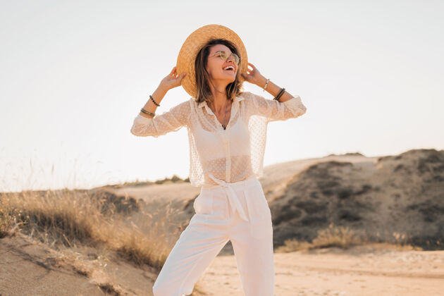 阳光时尚快乐的美丽微笑的女人在沙漠里摆姿势 穿着白色的衣服 戴着草帽和太阳镜在夕阳下休闲旅游沙丘