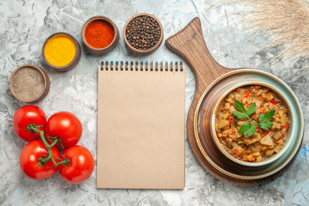 碗砧板上碗里烤茄子沙拉和碗里不同香料的俯视图灰色表面上的一个笔记本西红柿香料茄子沙拉蔬菜