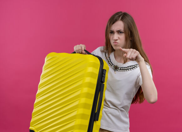 手提箱恼怒的年轻旅行家女孩拿着手提箱 指着孤立的粉红色空间的左侧年轻的旅行者女孩